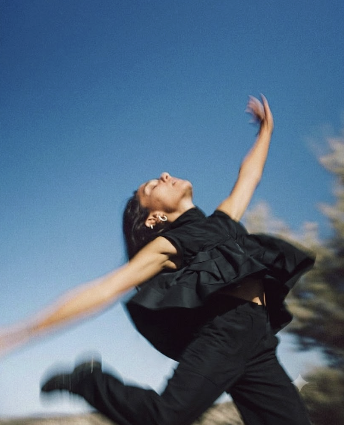 Woman in white technical outdoor clothing leaping mid-air with flowing yellow ribbons against a bright blue sky